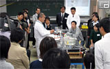理科実験土曜塾で、実験の説明をする本校教諭と受講者の皆さんの写真です。