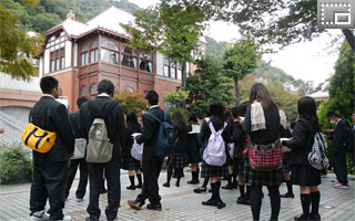クラス別行動で、神戸・北野異人館街散策に行く前、風見鶏の館前（北野町広場）で説明を受ける生徒たちの写真です。