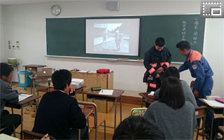 職業人講話－講師（消防士）の指示で消防服を着る生徒と、その様子を見守る生徒たちの写真です。